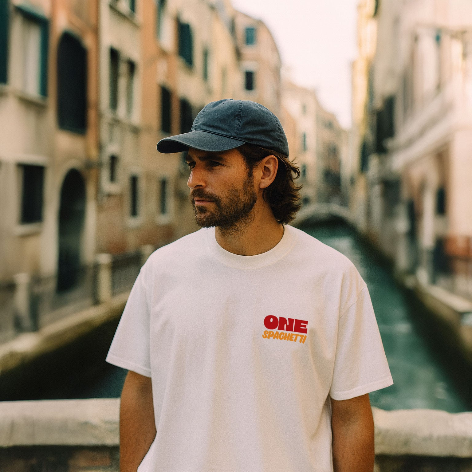 Young man wearing a cap and oversized white One Spaghetti Chef’s Choice T-shirt with front print, standing in Venice. Limited edition streetwear.